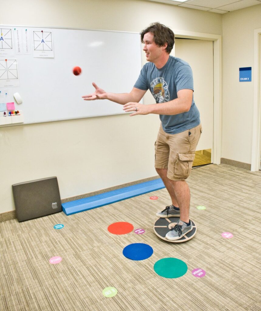 A patient during vestibular system and vision rehabilitation exercise balancing on a Bosu ball while playing catch and a memory game.