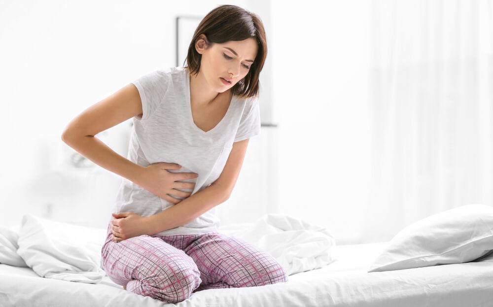 A woman is sitting on her bed grabbing her stomach in pain.