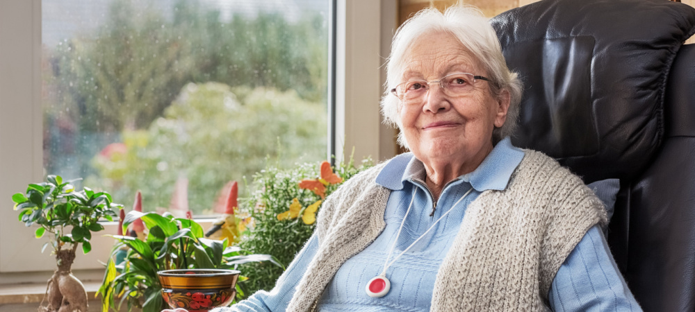 An elderly woman sits near window with an emergency button near by. 